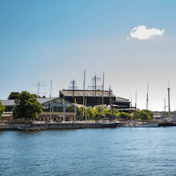 Hamnscen med olika segelbåtar dockade längs en strandpromenad, stor byggnad märkt "Junibacken", träd och en klar himmel.