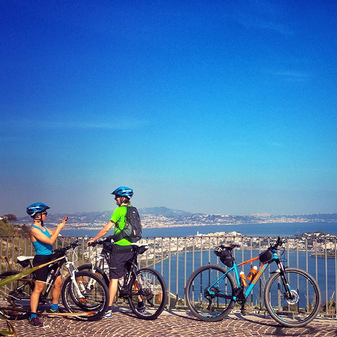 Due ciclisti con il casco si fermano ad ammirare il panorama della città da un belvedere. Due biciclette accanto a loro, cielo e acqua sullo sfondo.