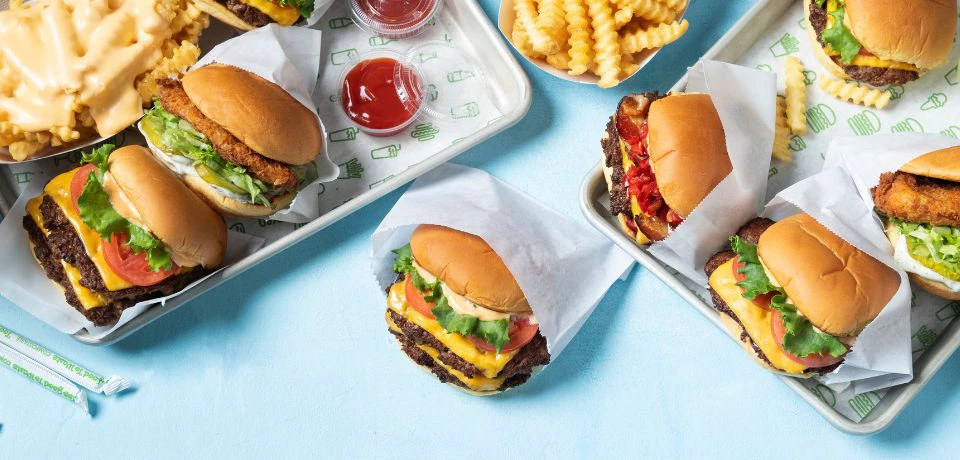 Assorted burgers in wrappers, crinkle-cut fries, and ketchup in small containers on a light blue background.