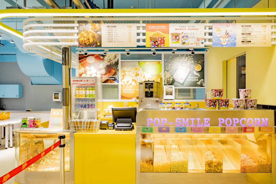 A colorful popcorn stand with a yellow counter, popcorn bins, a register, and a menu above advertising various options.