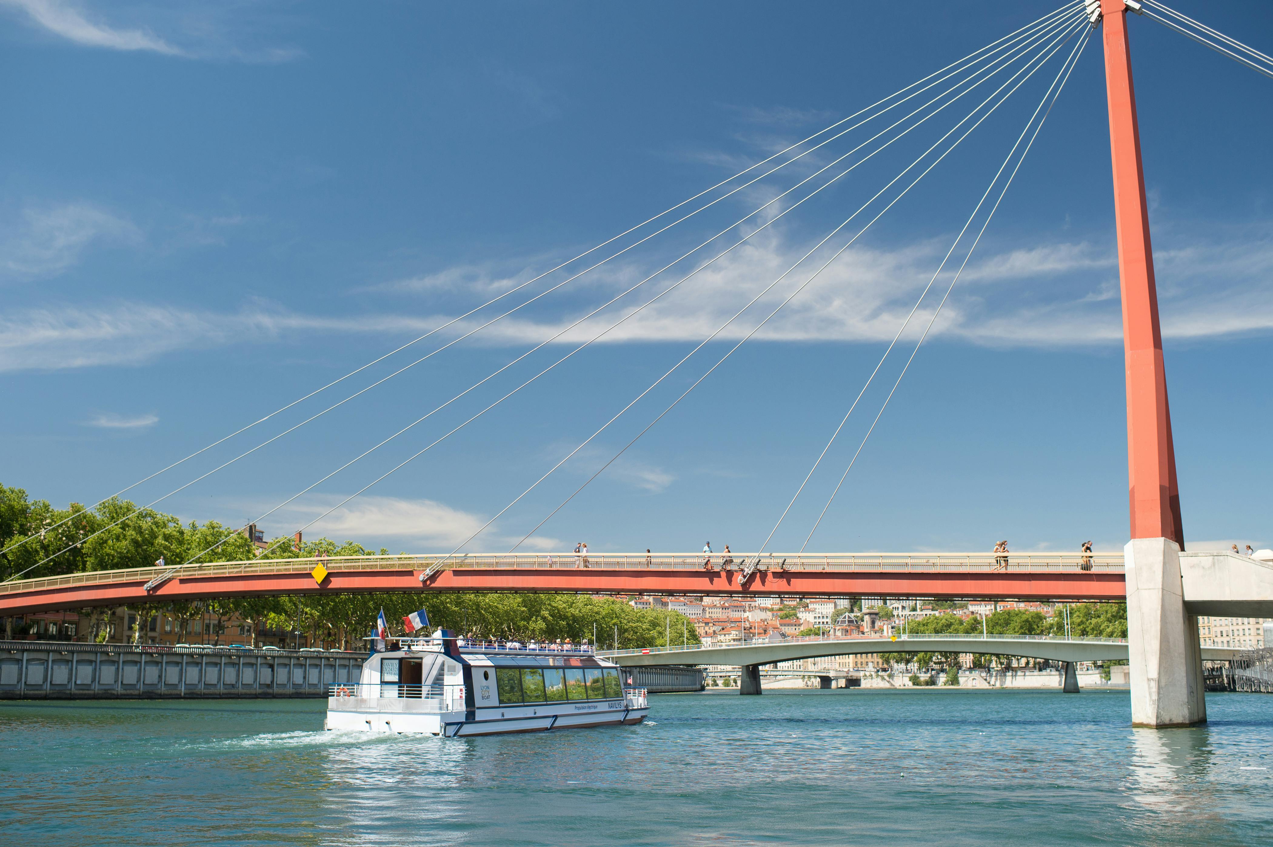 A white boat floats on a river under a suspension bridge with cables, against a backdrop of buildings and trees under a blue sky.