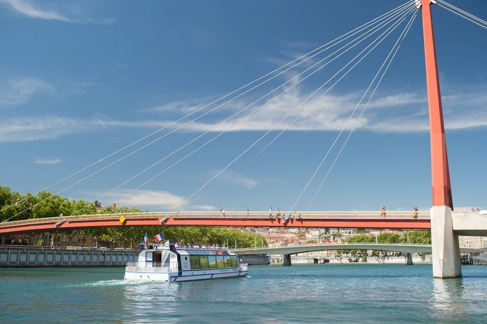 A white boat floats on a river under a suspension bridge with cables, against a backdrop of buildings and trees under a blue sky.