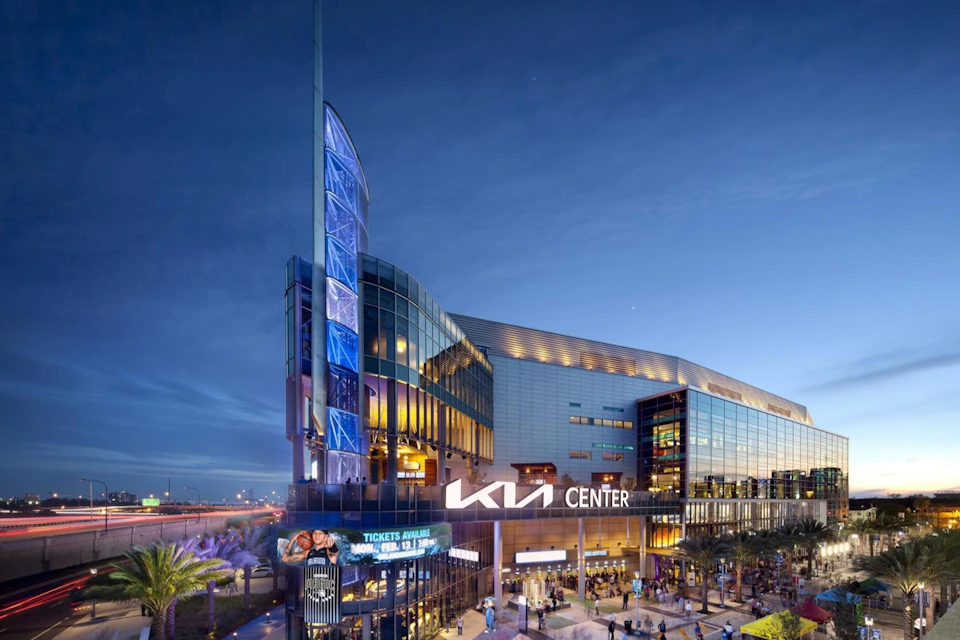 Modern building with "KIA Center" signage, lit exterior with large blue screen, bustling crowd, and evening sky in the background.