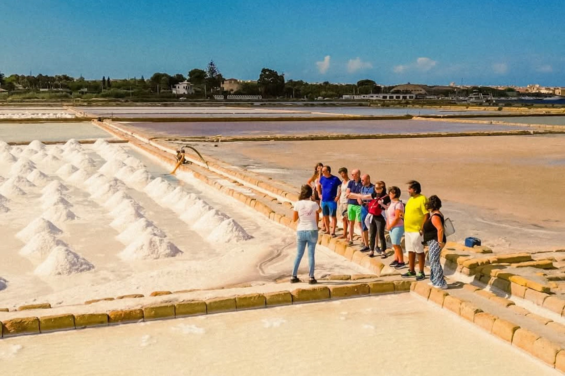 A group of people standing near white salt mounds in a salt flat area under a clear sky, with distant buildings visible.