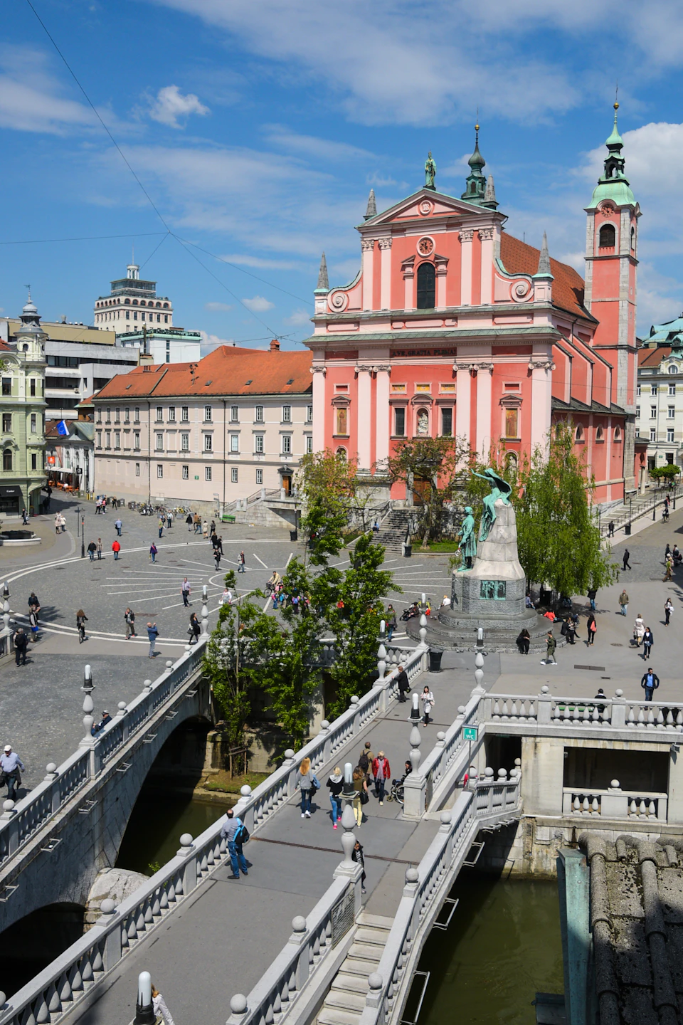 A bustling city square with people walking, a pink baroque-style church, a bridge, and a statue in the center.