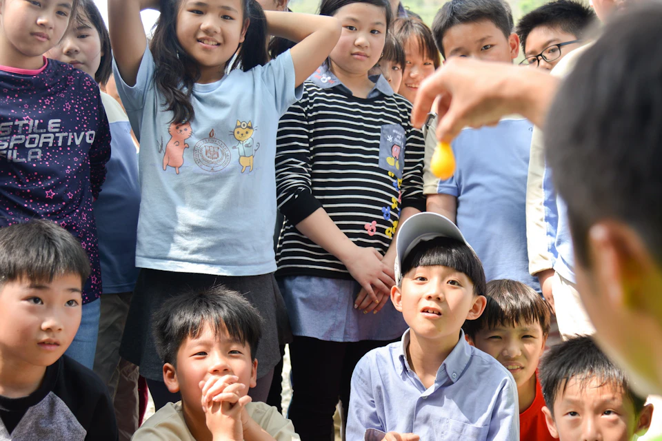 A group of children outdoors observing someone holding a small object in focus at the front of the scene.