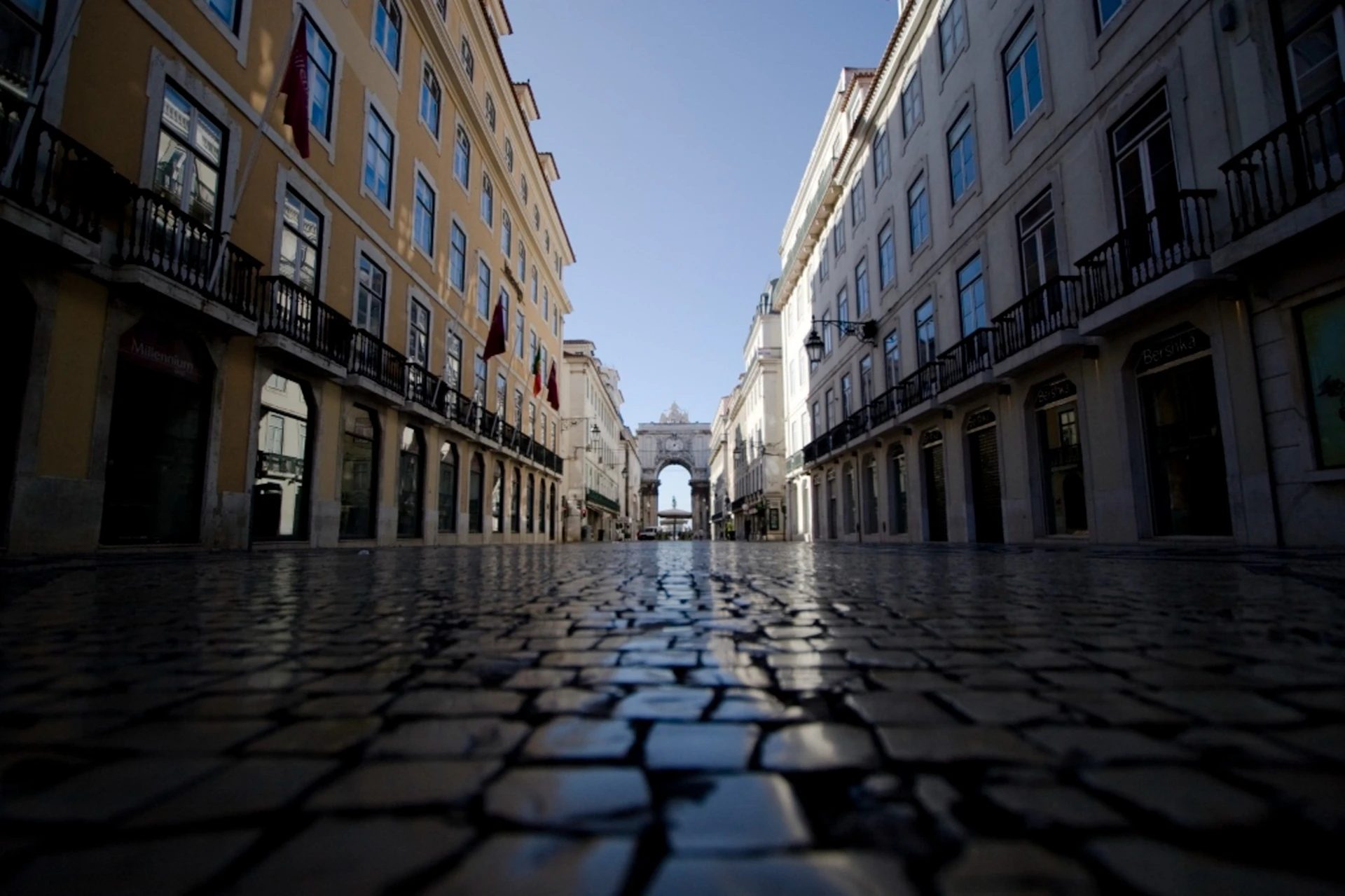 Low angle view of a cobblestone street flanked by buildings with balconies, leading to an archway under a clear blue sky.