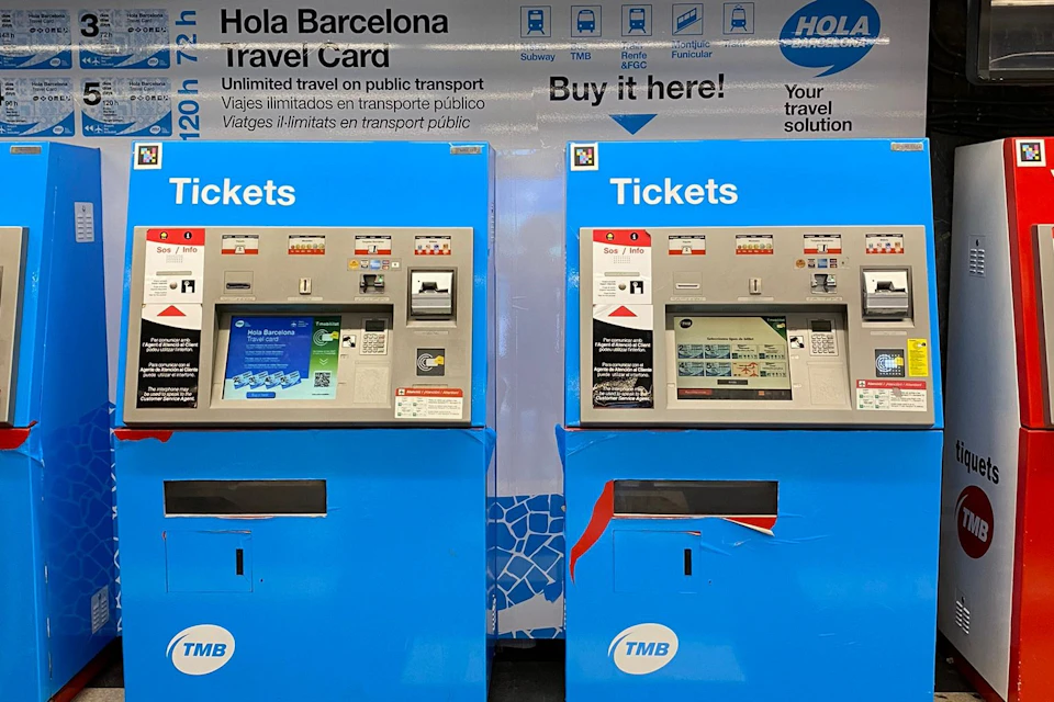 Two blue ticket vending machines with screens and slots, marked "Tickets," under a sign advertising Hola Barcelona travel cards.