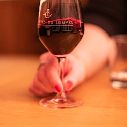 A hand holding a wine glass with red wine, placed on a wooden table. The glass has text "Caves du Louvre" on it.
