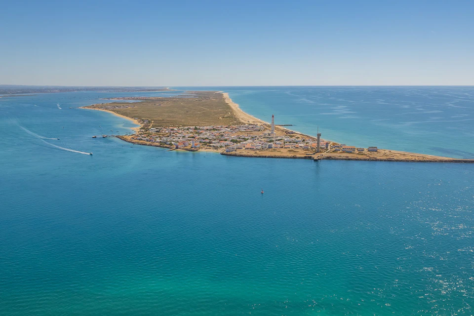 Aerial view of Farol Island with lighthouses, sandy beaches, and blue ocean stretching along a narrow peninsula.
