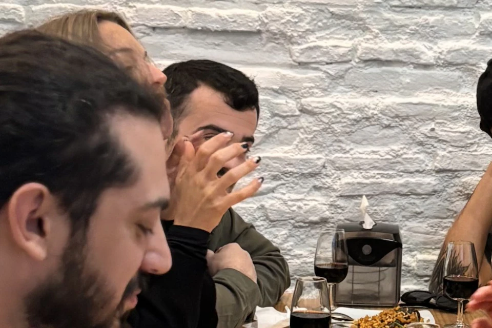 A group of people sits around a wooden table, eating food from white plates and drinking dark beverages.