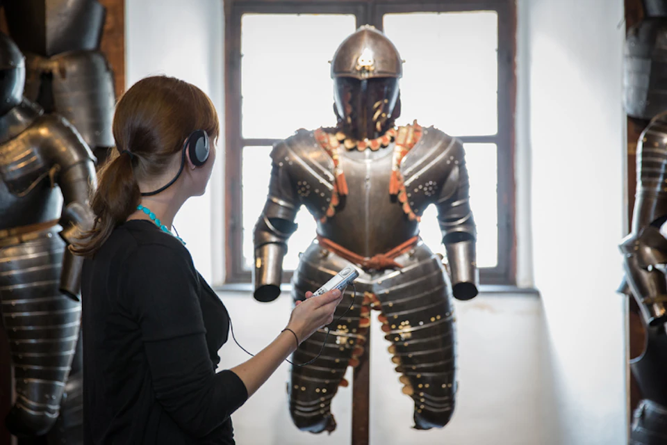 Woman with headphones and audio guide looking at a suit of armor displayed in front of a large window.