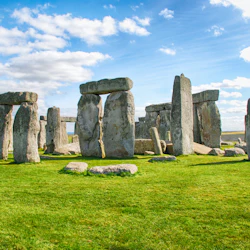 Stonehenge med stora stående stenar arrangerade i en cirkel på ett grönt gräsbevuxet fält under en blå himmel med spridda moln.