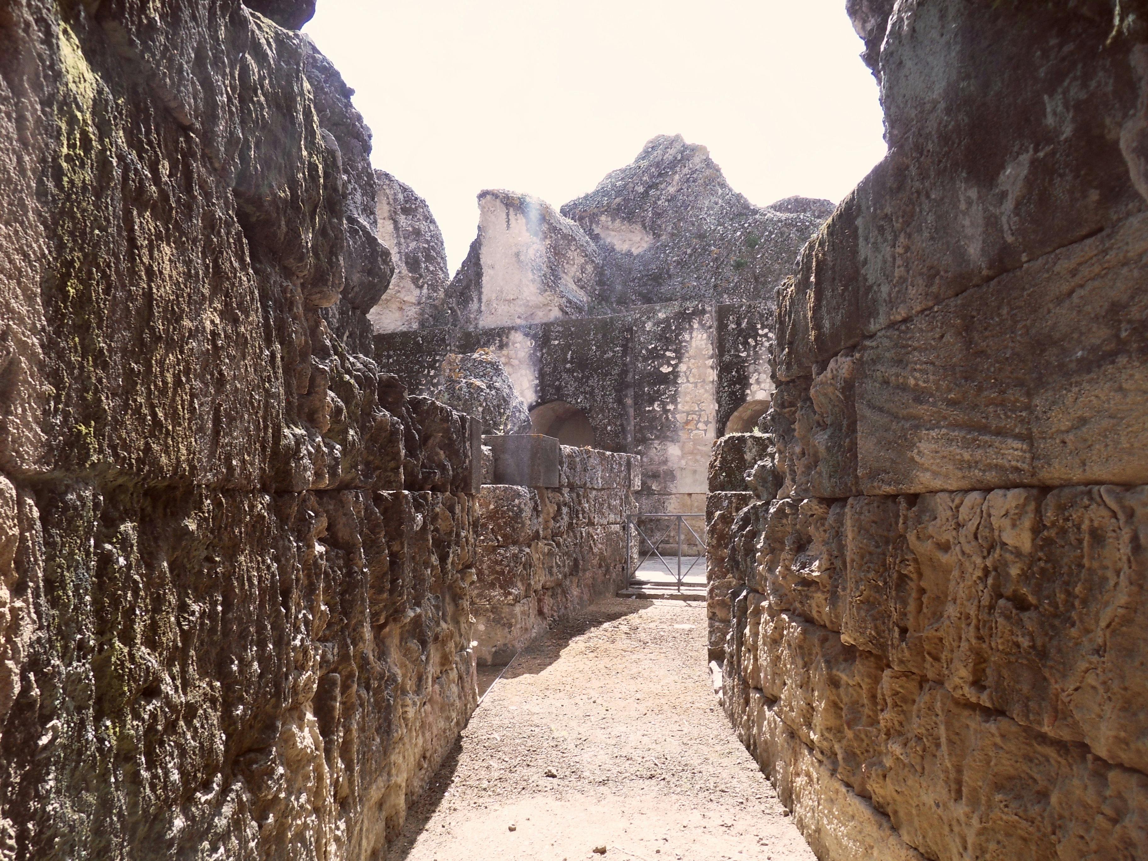 Cesta mezi starobylými kamennými zříceninami se zvětralými, rozpadajícími se zdmi vedoucí k částečně uzavřenému prostoru. Jasná, sluncem zalitá scéna.
