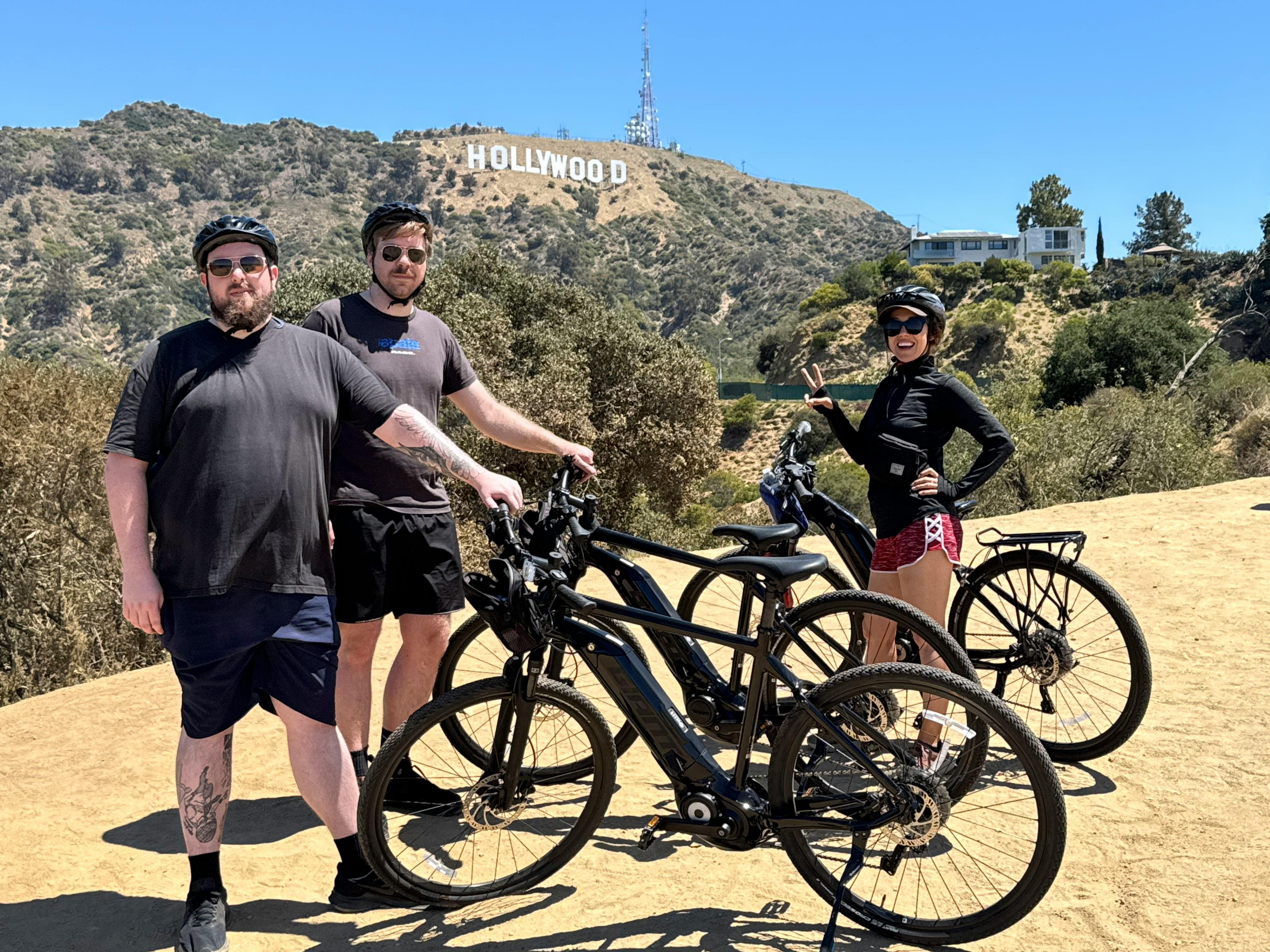 Three people pose with bicycles in front of the Hollywood sign on a sunny day.