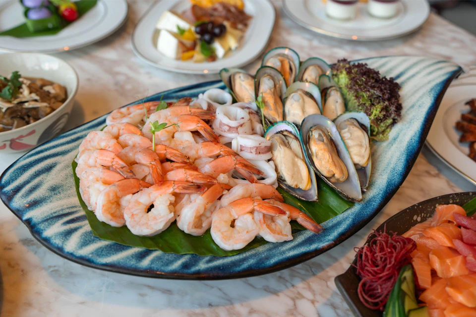 A seafood platter with shrimp, squid rings, green mussels, and greens on a blue plate, with more dishes in the background.