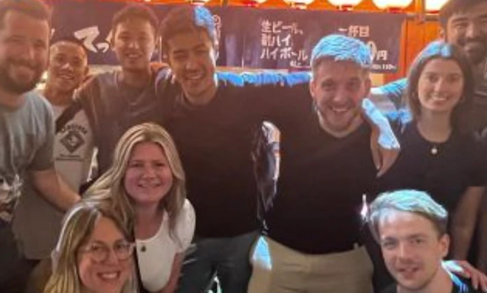 A group of people smiling and posing outside a lit-up establishment with Japanese signs and lanterns at night.