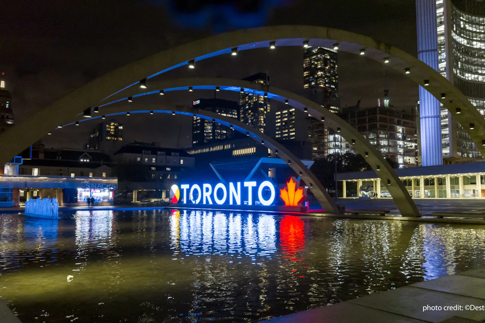 The Toronto sign at Nathan Philips Square lit up at night and framed by the concrete arches above the water feature