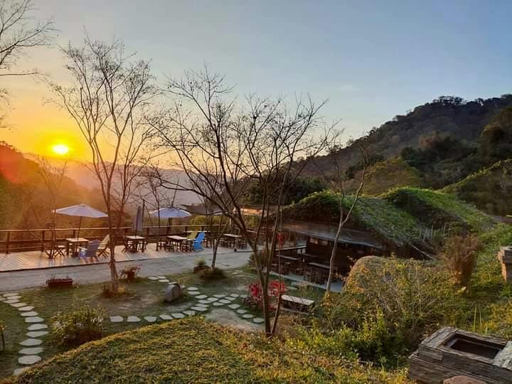 A scenic outdoor café with tables and umbrellas, surrounded by greenery and trees, during sunset near a hillside.