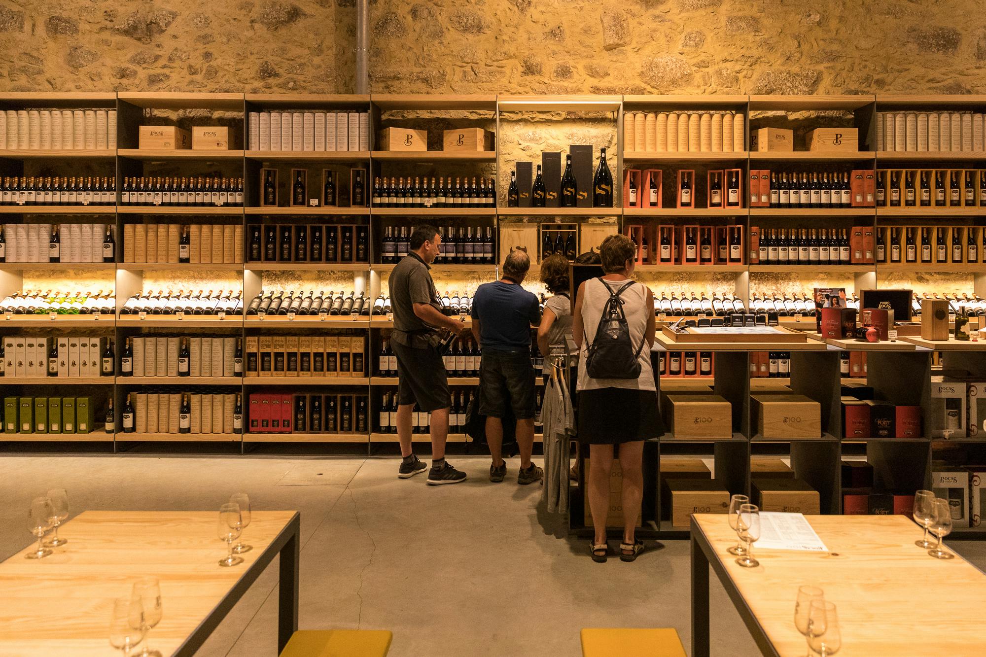 石の壁を覆う木製の棚の上にあるワインボトルを調べているワインショップに4人が立っています。 メガネの空いたテーブルが目の前にあります。