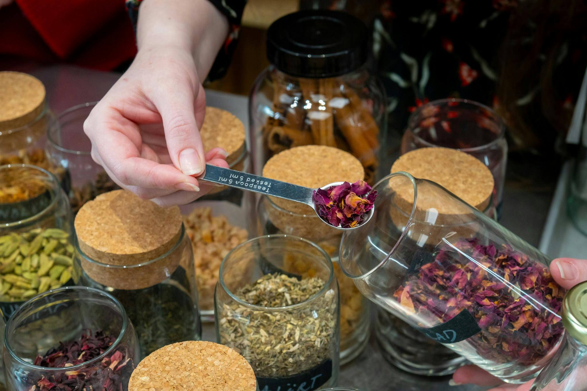 Une main tenant une cuillère à café avec des pétales de rose séchés au-dessus de bocaux en verre contenant des herbes et des épices, certains avec des couvercles en liège.