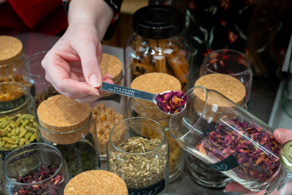 A hand holding a teaspoon with dried rose petals over glass jars of herbs and spices, some with cork lids.