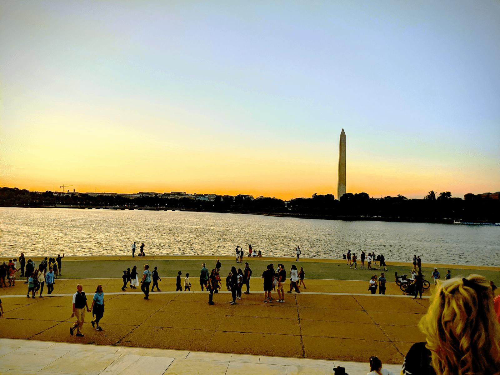 워싱턴 기념비를 배경으로 석양에 반사되는 커다란 수영장 근처를 거닐고 있는 사람들.