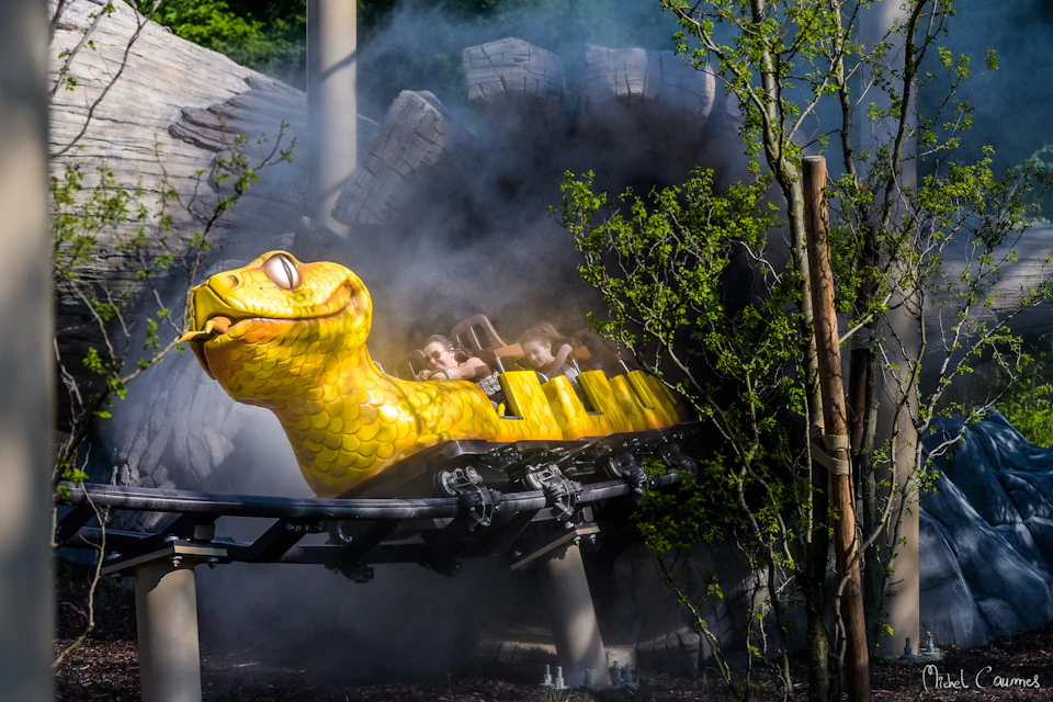 Rollercoaster with a bright yellow snake head car emerging from smoke, with riders visible and trees surrounding the track.
