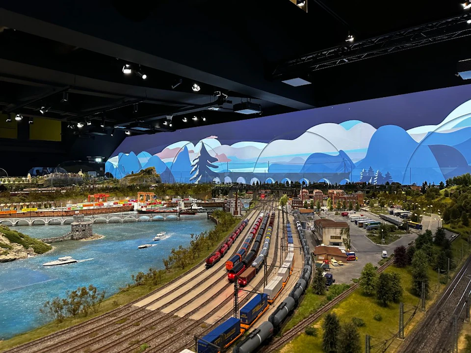 A detailed model train layout featuring tracks, trains, buildings, a river, and a scenic backdrop mural of mountains and sky.