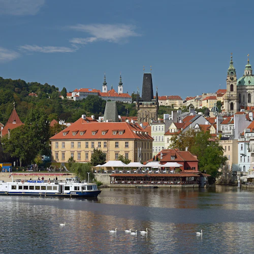 Flod med en båt märkt "PRAGBÅTAR", stadsbild med byggnader med röda tak, torn, svanar på vattnet och kullar i bakgrunden.