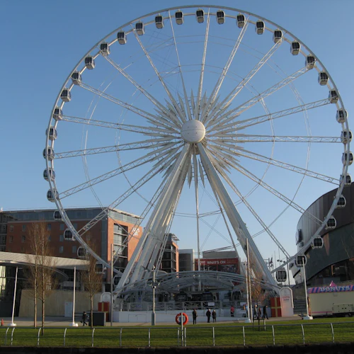 Stort pariserhjul framför moderna byggnader med en klarblå himmel, omgiven av grönt gräs och ett munkstativ.