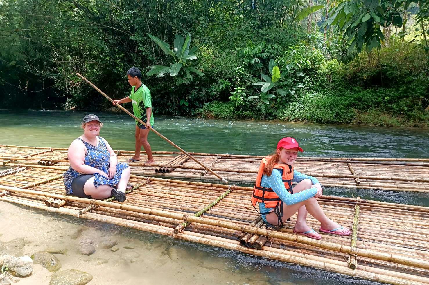 Two people sitting on a bamboo raft while a guide stands and steers with a pole on a river surrounded by lush greenery.