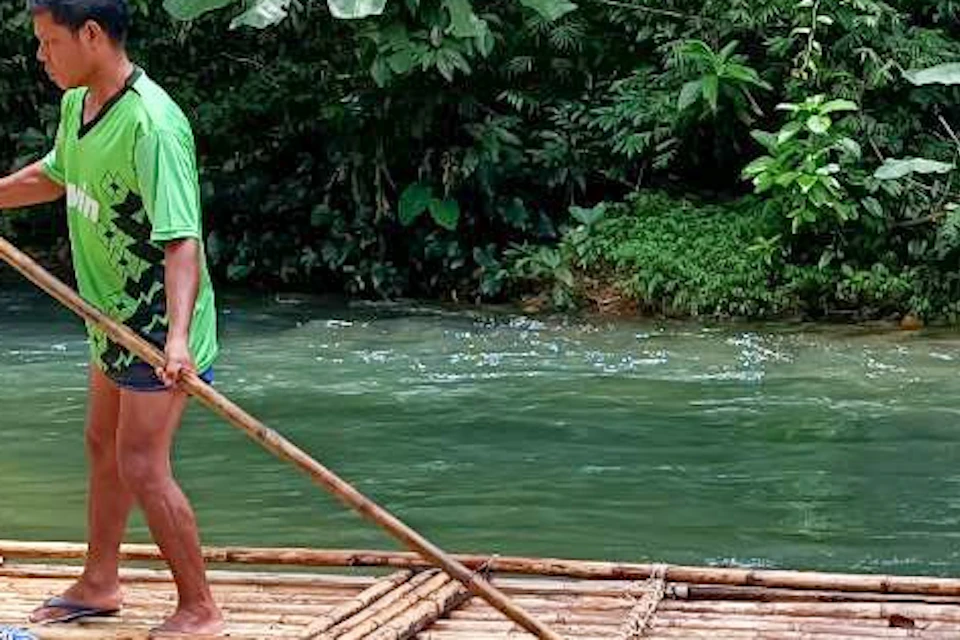Two people sitting on a bamboo raft while a guide stands and steers with a pole on a river surrounded by lush greenery.