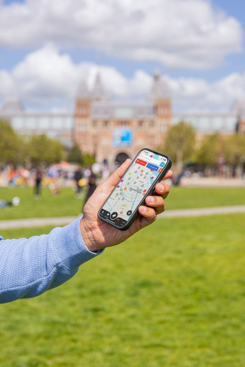 A person holding a smartphone with a map app open in a grassy park with a large building and people in the background.