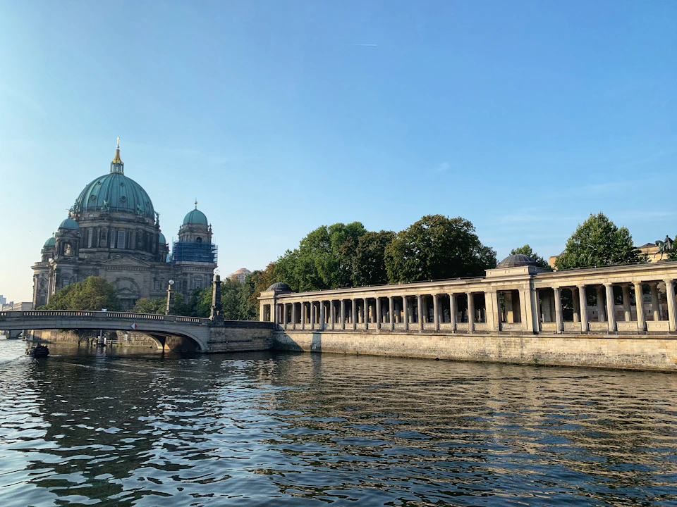 A domed cathedral and a colonnaded building flank a bridge over a calm river, with trees in the background under a clear blue sky.