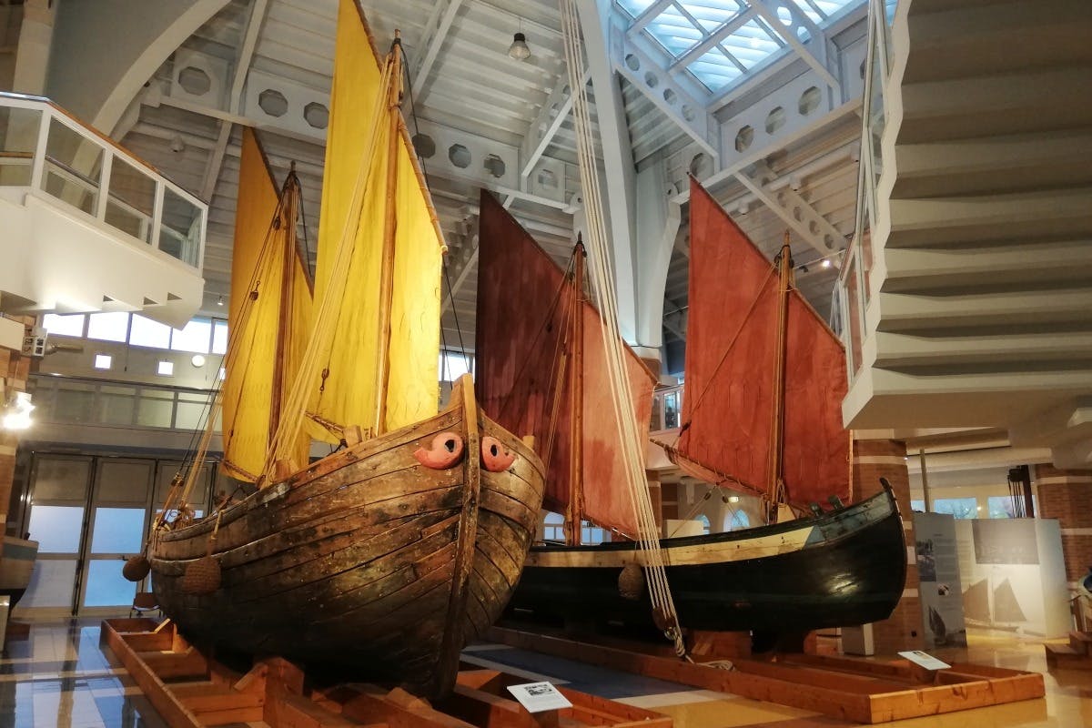 Due barche a vela in legno con vele gialle e rosse sono esposte all'interno sotto un moderno soffitto a travi metalliche.