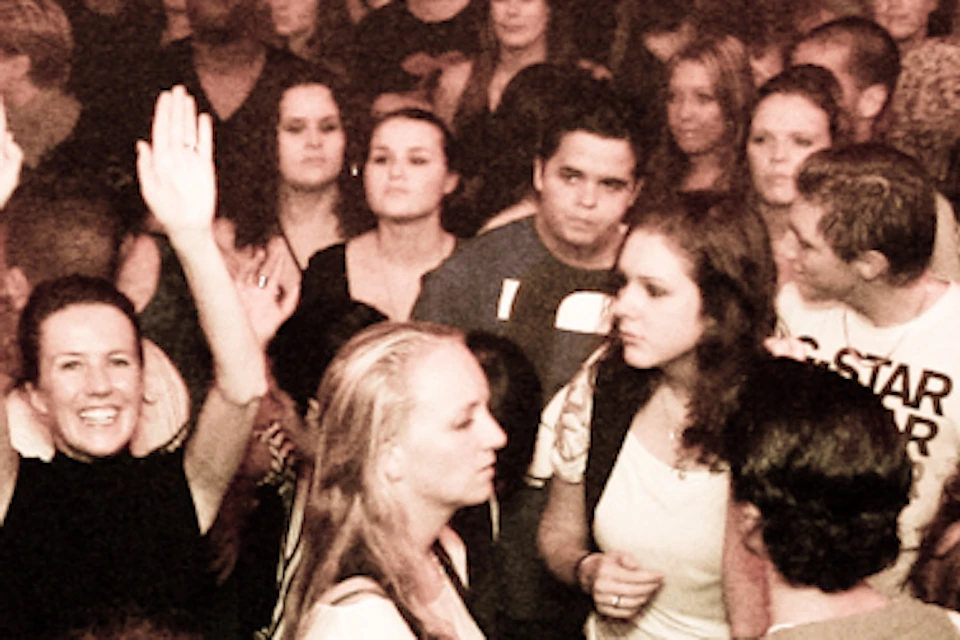 A DJ wearing headphones performs for a large, energetic crowd at a packed nightclub. The text reads "Amsterdam Nightlyfe."