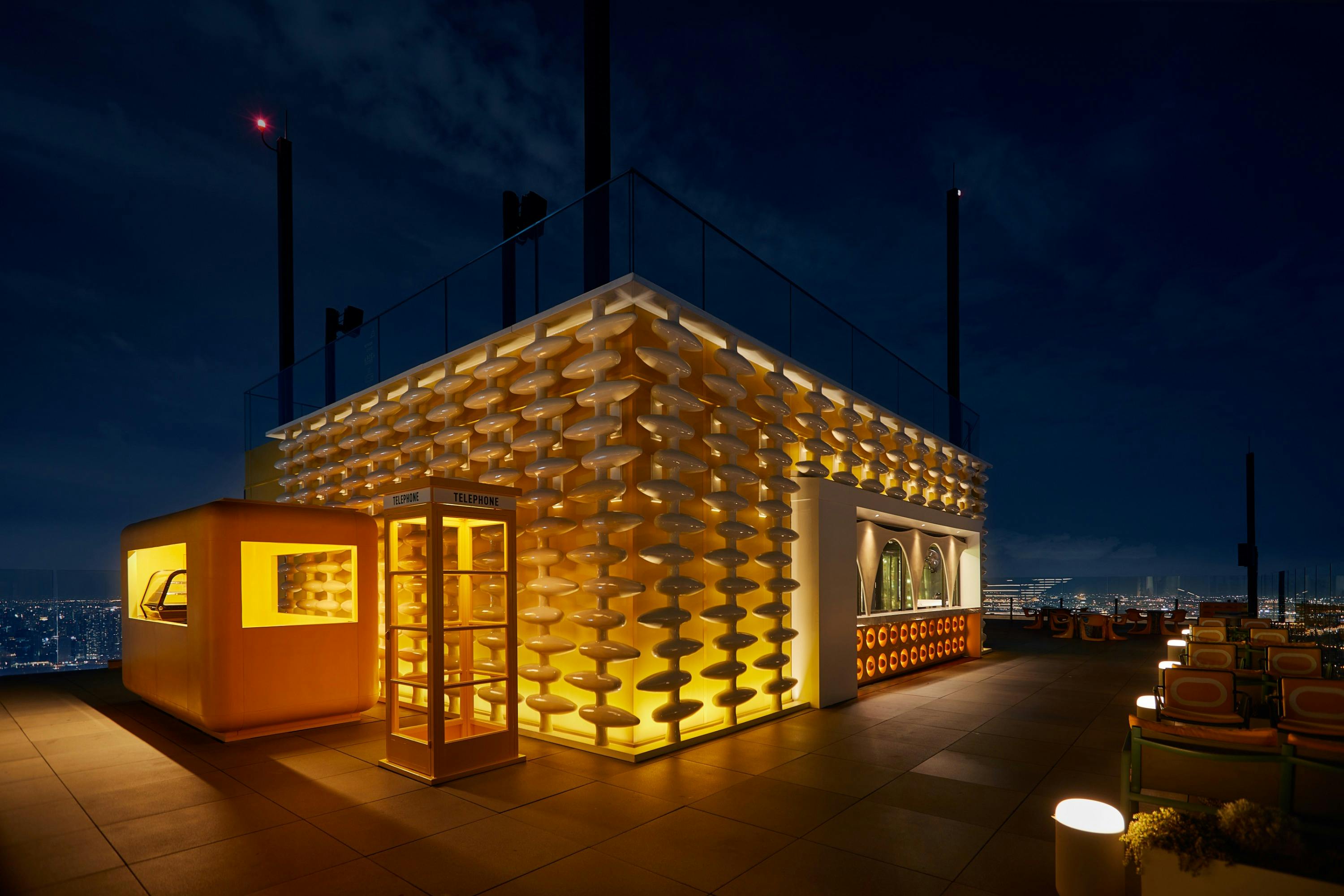A modern rooftop space at night with illuminated geometric walls, a glass booth, and city lights in the background.