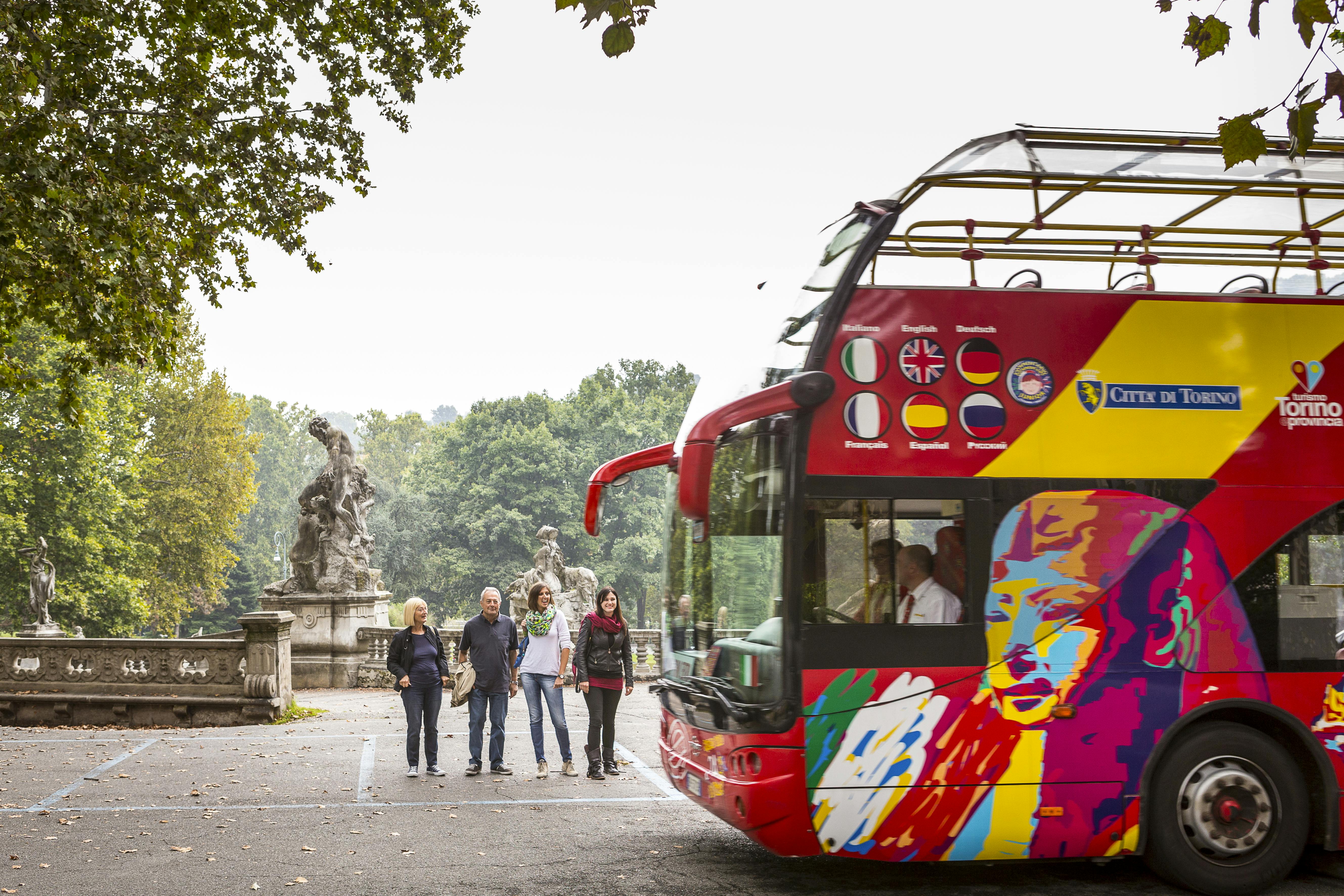 Čtyři lidé stojí u barevného dvoupatrového turistického autobusu zdobeného různými vlajkami, zaparkovaného u sochy v zeleném parku.