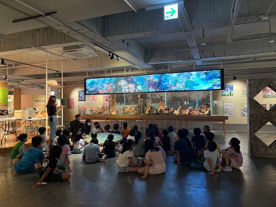 A group of children seated on the floor of an exhibit space, looking at colorful artwork displayed on a large screen and walls.
