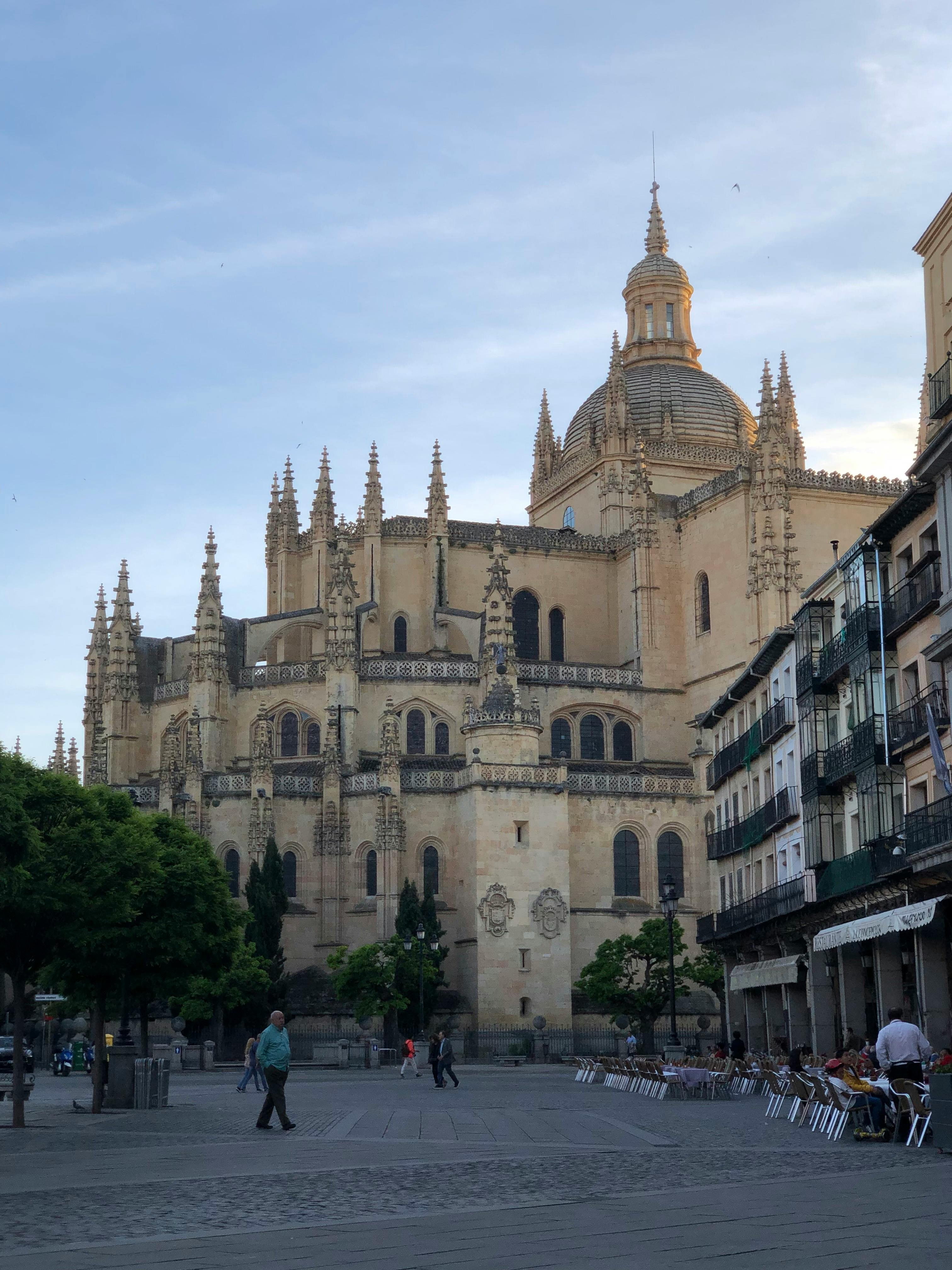 A cathedral with intricate gothic architecture dominates a public square, flanked by trees and buildings with outdoor seating.