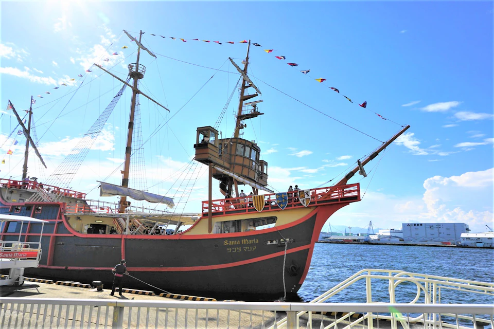 A replica of a historical sailing ship named 'Santa Maria' docked at a pier under a sunny sky, with flags on its masts.