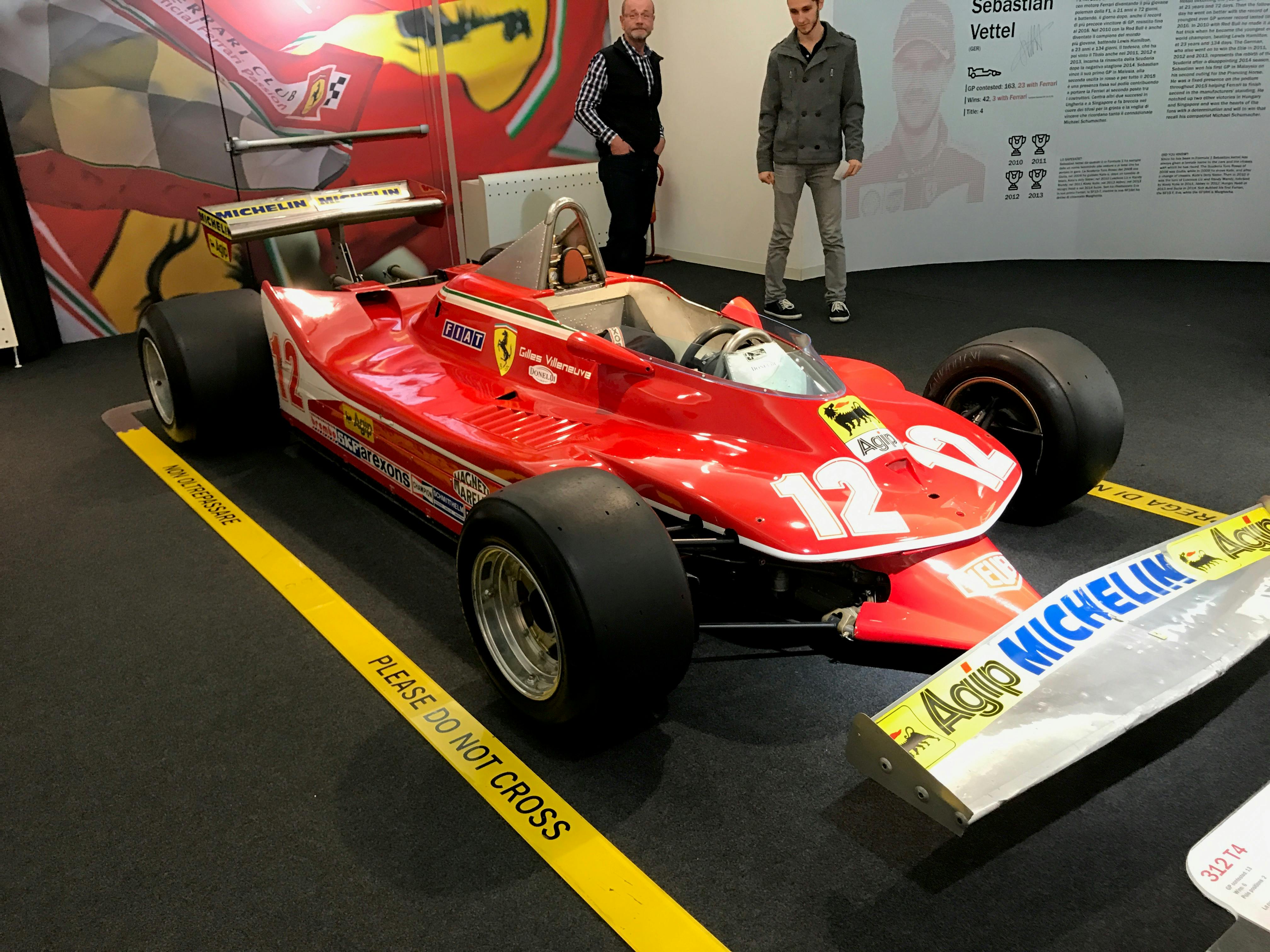 Κόκκινο αγωνιστικό αυτοκίνητο Formula 1 vintage με τον αριθμό 12 στο καπό, που εμφανίζεται σε εσωτερικό χώρο. Δύο άντρες στέκονται πίσω του.