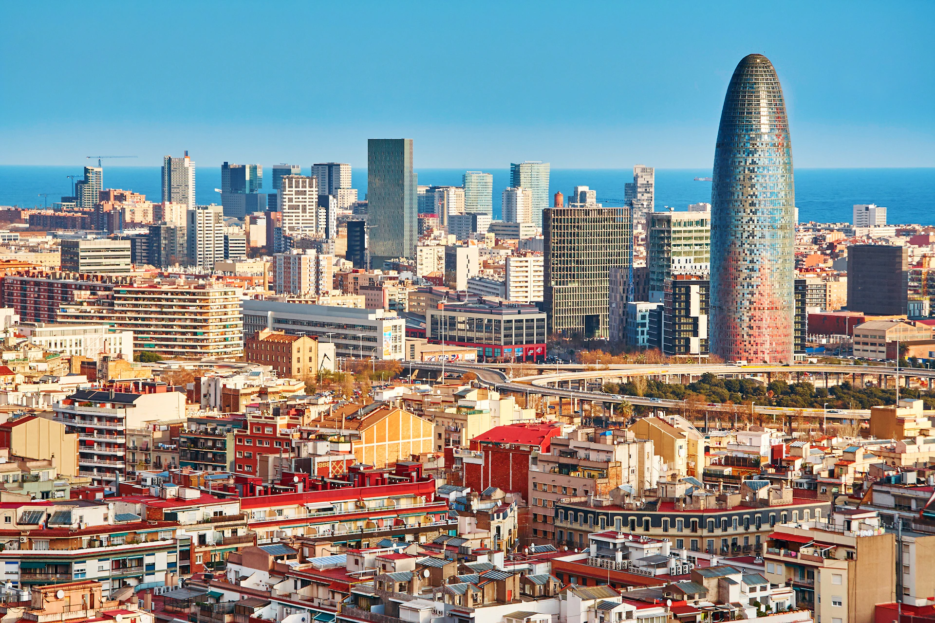 Cityscape with a mix of modern skyscrapers and older buildings, dominated by a tall, cylindrical, mosaic-like tower. Blue sky in the background.