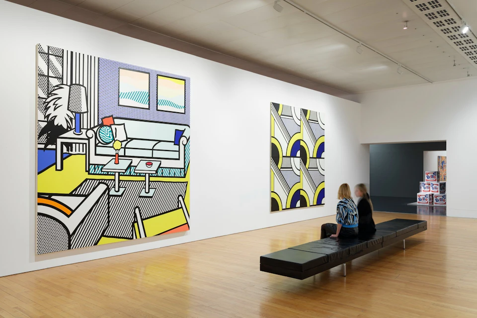 Two people sit on a bench viewing large colorful paintings in a modern art gallery with wooden floors and white walls.