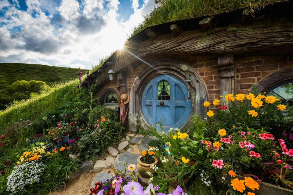 A cozy grass-roofed house with a blue round door, surrounded by colorful flowers under a bright sky with sunlight.