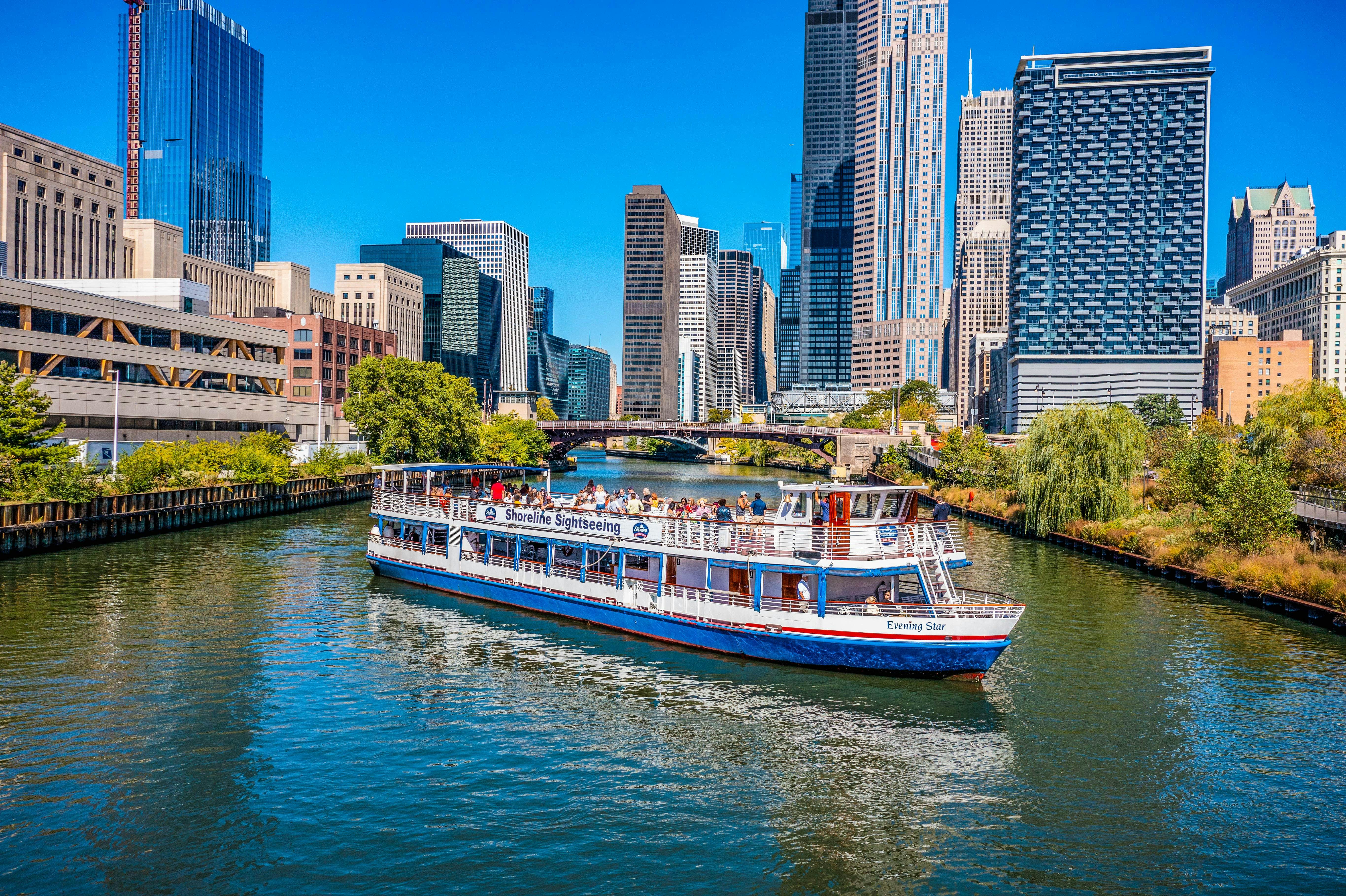 A sightseeing boat cruises along a river with modern skyscrapers in the background on a clear, sunny day.