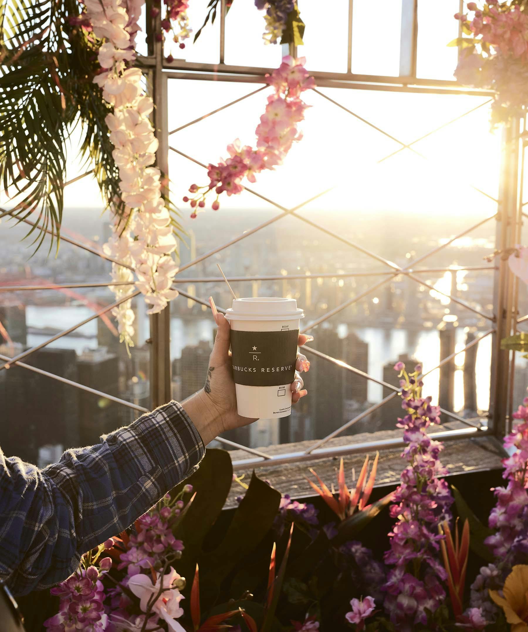 A hand holding a Starbucks cup with cityscape and sunset in the background, surrounded by pink and purple flowers.
