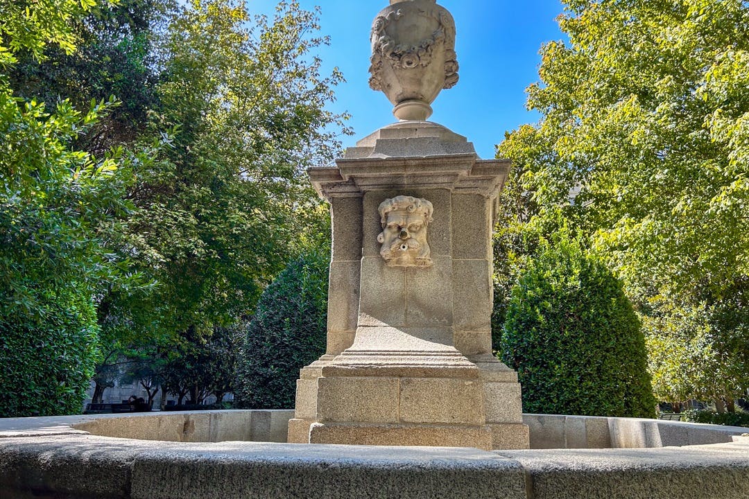 Μια πέτρινη βρύση με διακοσμητική τεφροδόχο και γλυπτό πρόσωπο, περιτριγυρισμένη από δέντρα και πράσινο κάτω από έναν καταγάλανο ουρανό.