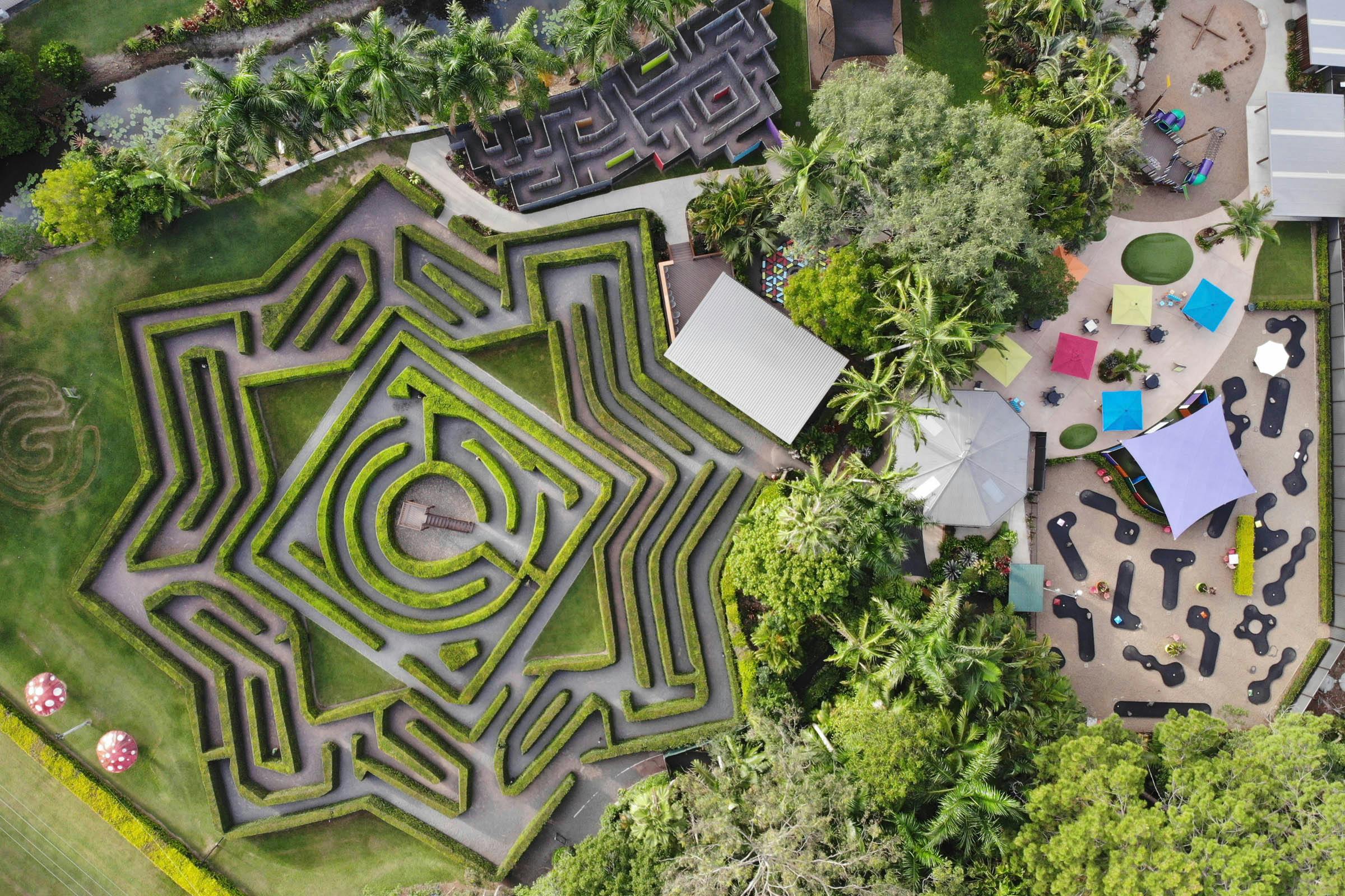 Вид с воздуха на геометрический лабиринт изгороди, детскую площадку с красочными зонтиками и окружающие деревья и дорожки.
